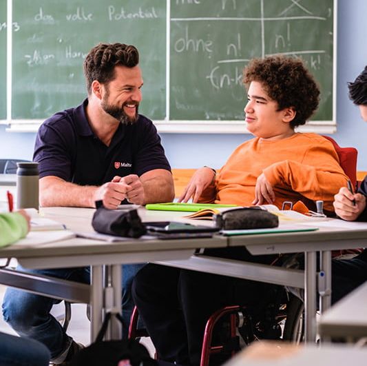 Malteser Schulbegleiter sitzt mit einem Jugendlichen im Rollstuhl im Klassenraum.