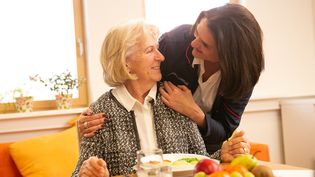 Malteser Mitarbeiterin beugt sich freundlich zu älterer Frau runter, die an einem gedeckten Tisch sitzt