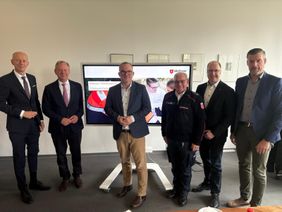 Sechs Männer stehen nebeneinander, im Hintergrund ein Bildschirm