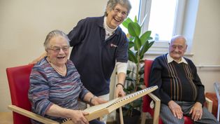 Eine Pflegekraft lacht fröhlich mit zwei älteren Menschen, die in bequemen Sesseln sitzen, eine Seniorin hält ein Holzspiel‑ oder Klanggerät in der Hand, im Hintergrund eine große Fensterfläche und eine grüne Pflanze sichtbar.