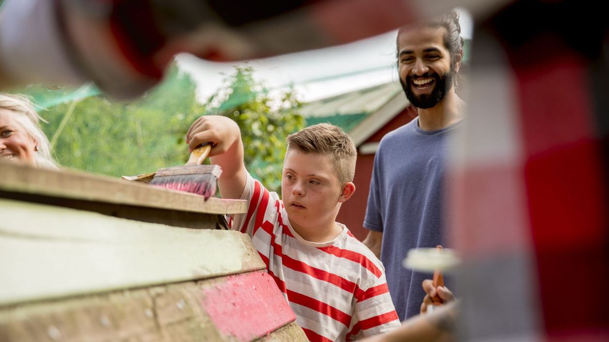 Inklusion in Deutschland: Junge streicht mit einem Betreuer eine Holzhütte an