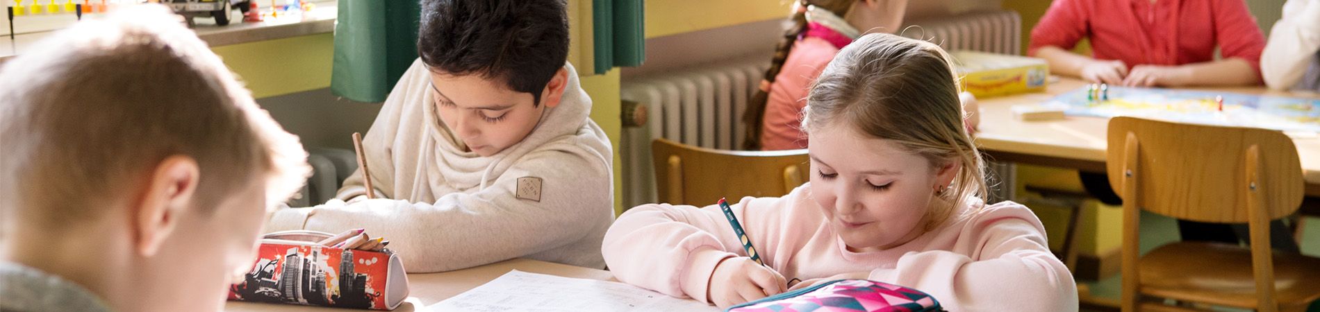 Drei Schüler sitzen an einem Tisch und schreiben. Zwei Jungen und ein Mädchen sind zu sehen. Sie befinden sich in einer Schulsituation und scheinen konzentriert zu arbeiten. Einer der Jungen hat schwarze Haare und trägt ein graues Sweetshirt, der andere Junge hat blonde Haare und ist nur von hinten zu sehen. Das Mädchen hat lange Haare, die zu einem Zopf zusammengefasst sind und trägt einen rosafarbenen Pulli. Auf dem Tisch sind Schulmaterialien und eine Federmappe zu sehen.