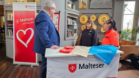 Ein Mann unterhält sich mit zwei Frauen. Sie stehen an einer Theke, auf der das Wort "Malteser" steht.