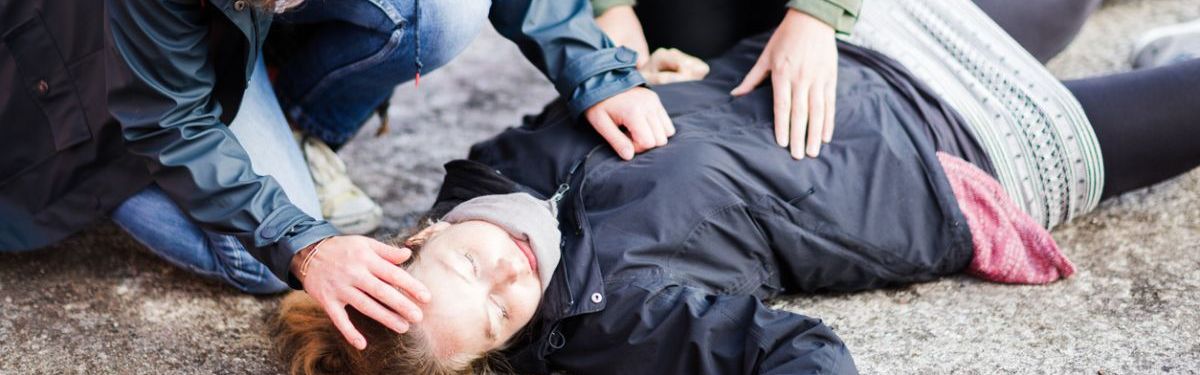 Zwei Frauen knieen auf einer Asphaltstraße über einer bewusstlosen Frau.