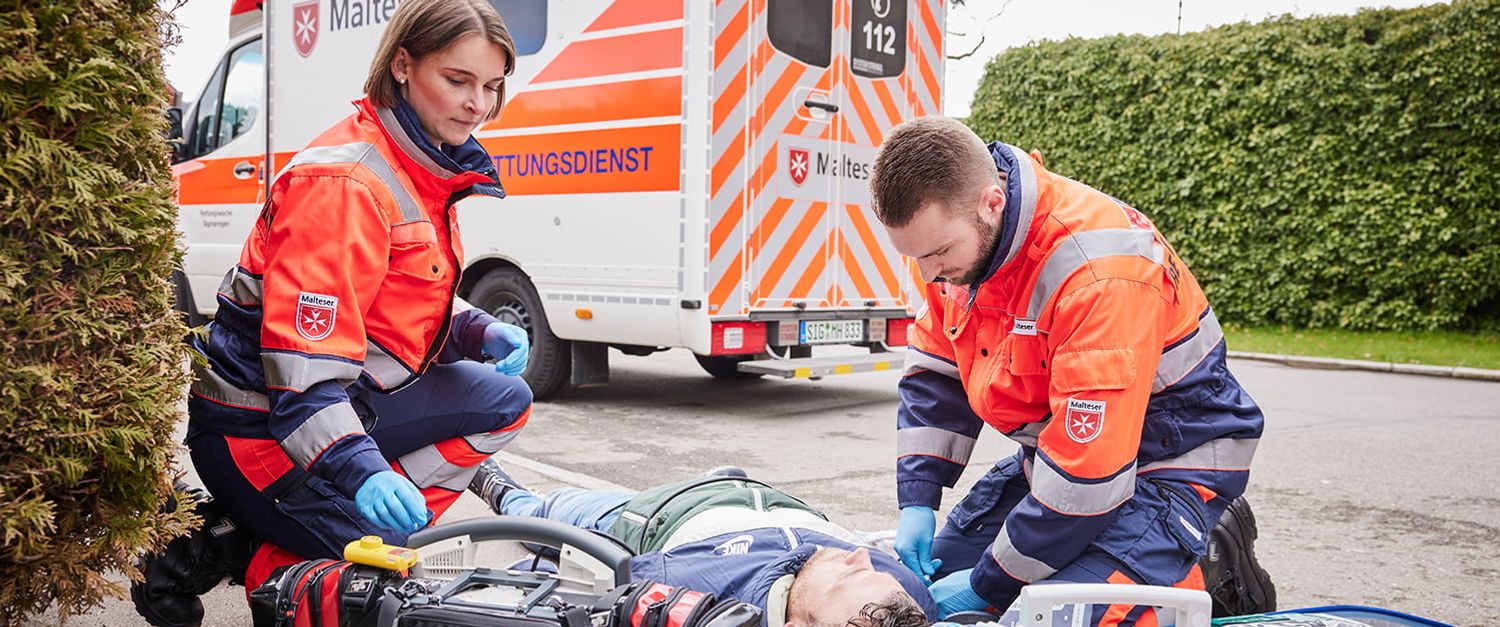 Eine Rettungssanitäterin und ein Rettungssanitäter versorgen einen Verletzten, der auf der Straße liegt. 