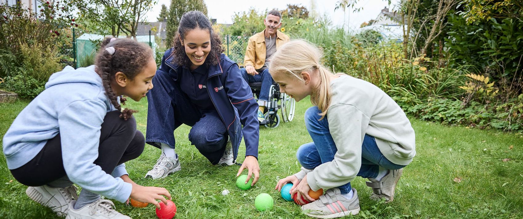 Frau mit Malteser Shirt spielt mit Kindern im Garten. 