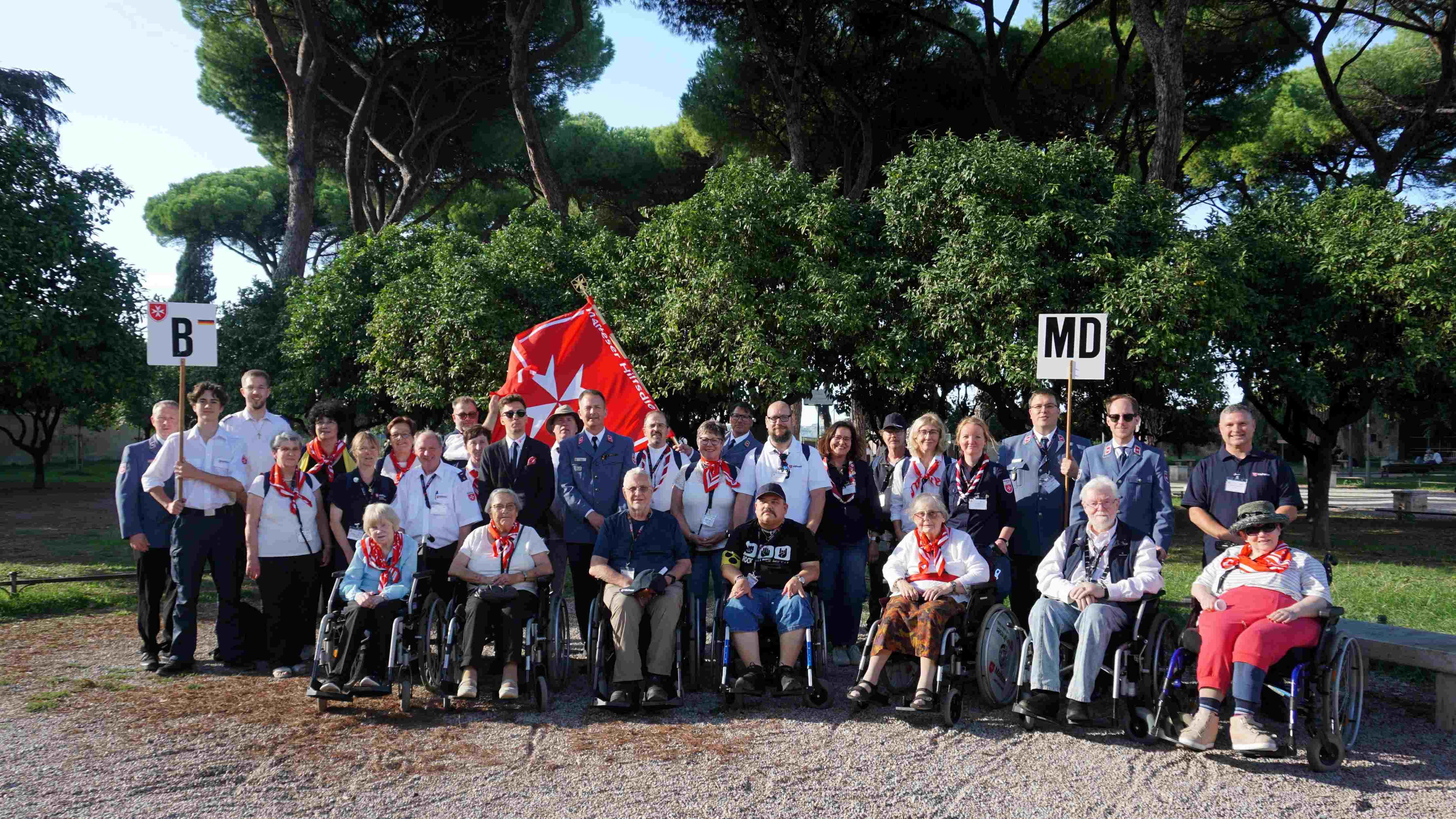Eine Gruppe steht zusammen in einem Park .Im Hintergrund schwingt eine rote Fahne mit weißen Malteser Kreuz.
