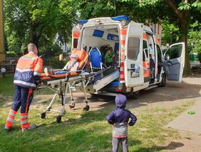 Ein Kind liegt auf einer Transportliege vor einem Rettungswagen. Andere Kinder schauen zu.