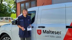 Sabine Meyer von den Rosenheimer Maltesern steht vor einem Malteser Auto und hält ein Hausnotrufgerät in den Händen.