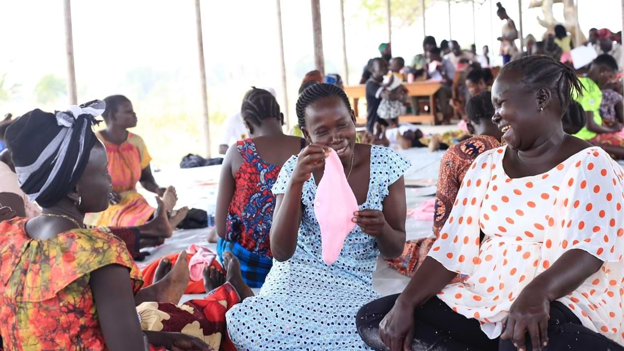 Eine Gruppe von Frauen in bunten Kleidern in einer Geflüchtetensiedlung in Uganda sieht sich eine Stoffbinde an und lacht.