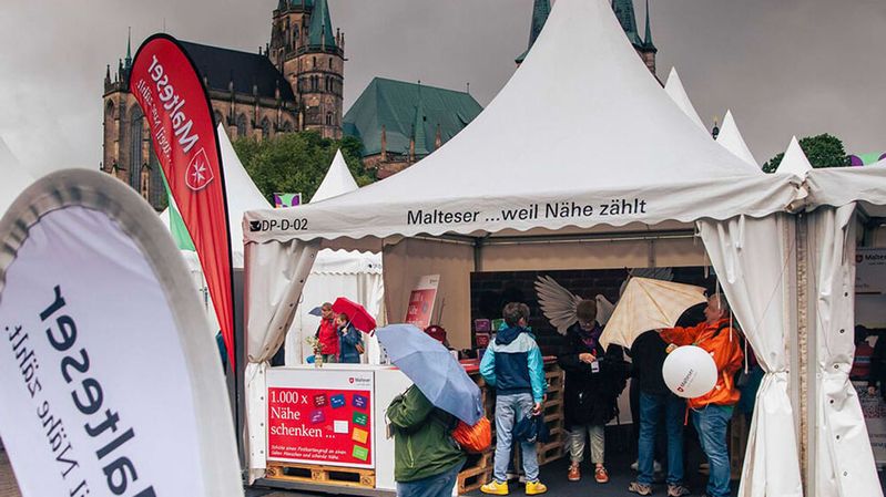 Mehrere Personen, teils mit Regenschirmen in der Hand, stehen unter einem weißen Zelt.