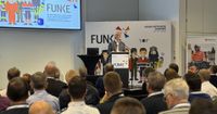 Ein Mann steht am Rednerpult, zahlreiche Menschen sitzen vor ihm