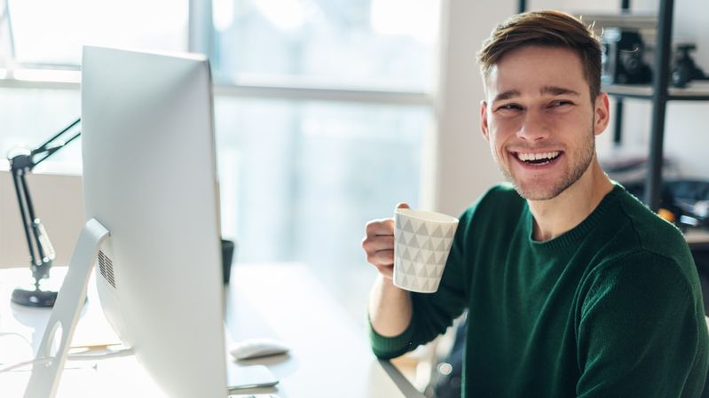 Junger Mann mit Kaffeetasse sitzt am Schreibtisch und lacht in die Kamera