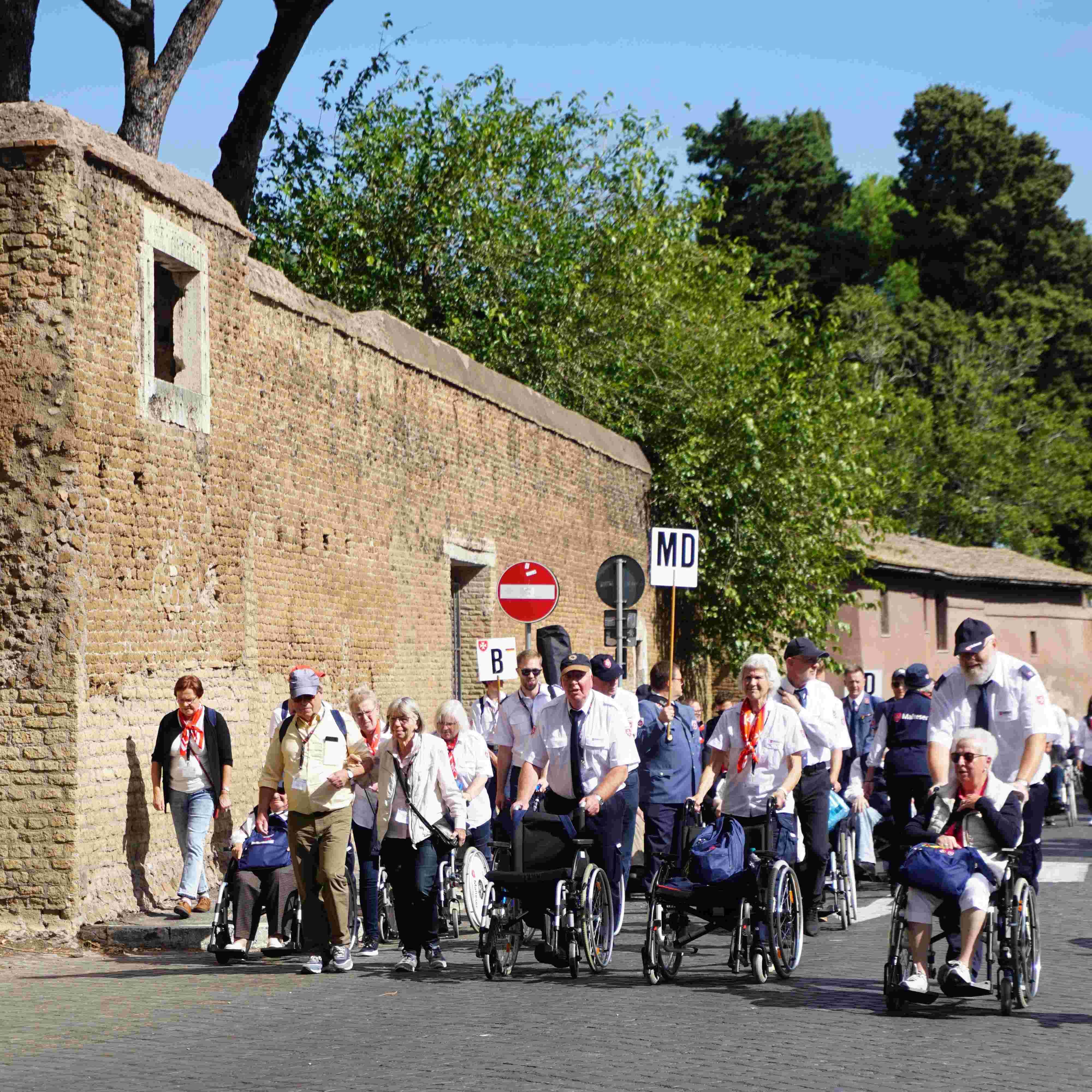 Gruppe von Menschen, darunter einige im Rollstuhl, geht auf einer Straße an einer hohen Steinmauer entlang, im Hintergrund Bäume und Verkehrsschilder.