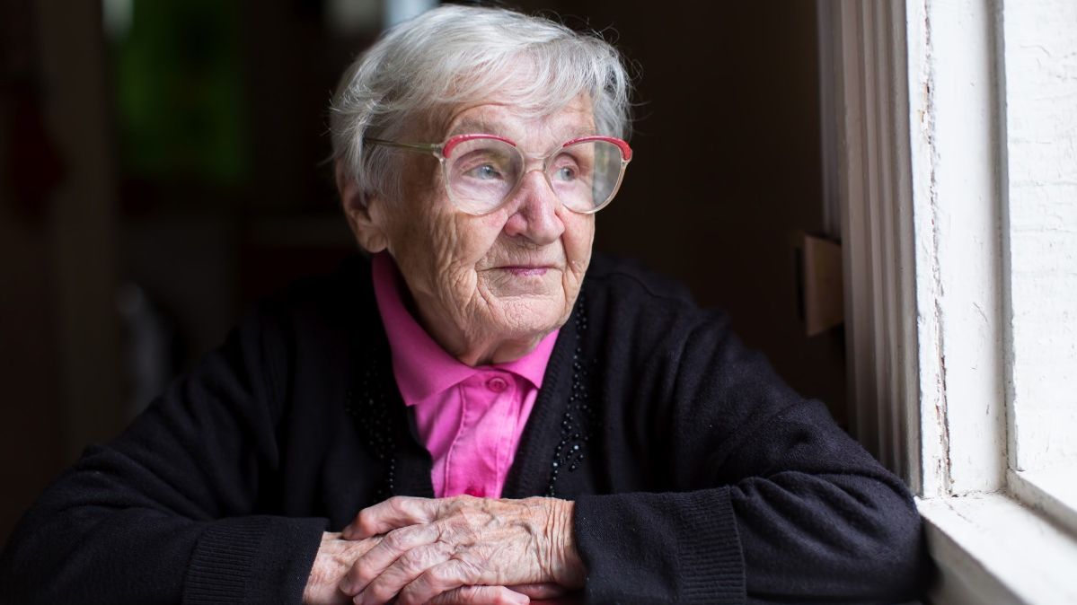 Eine alte Frau mit Brille schaut aus einem dunklen Raum aus einem Fenster und lächelt leicht.