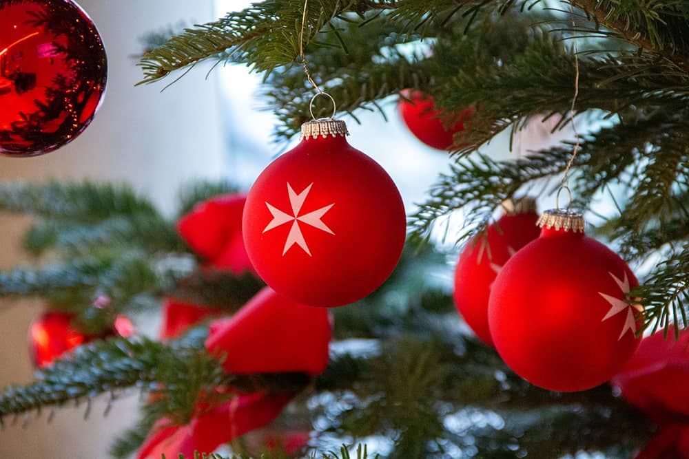 Rote Weihnachtskugeln mit dem achtspitzigen Wappenkreuz der Malteser hängen an einem Weihnachtsbaum. 