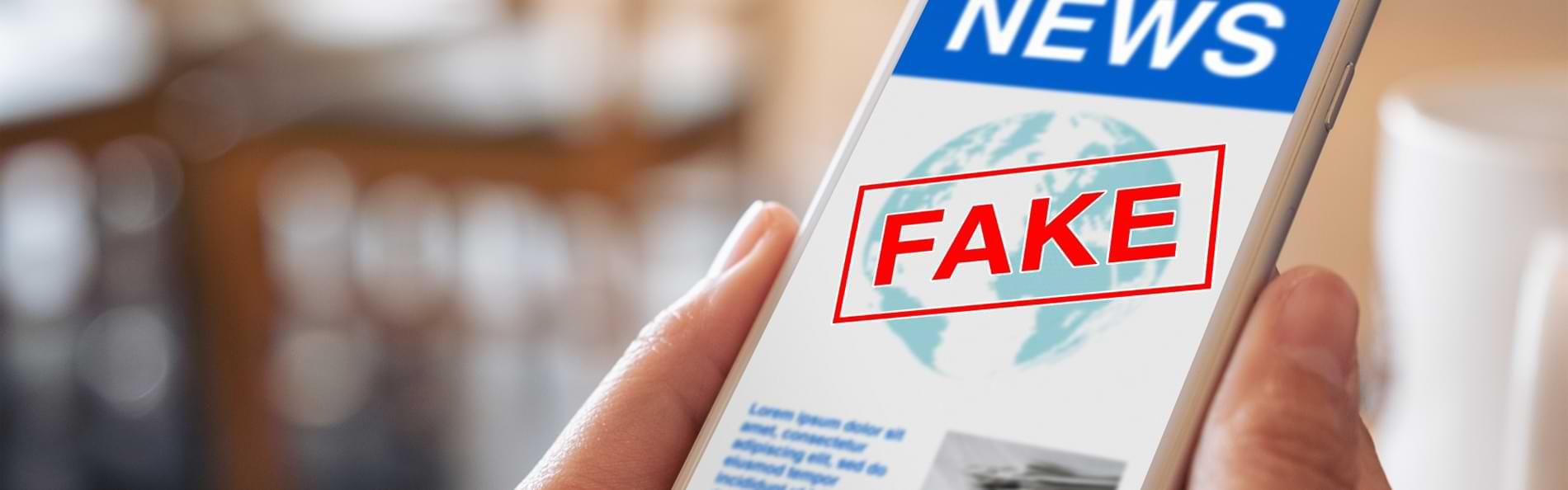 Zwei Hände halten ein Smartphone, auf welchem eine News-Seite zu sehen ist. Die Seite ist mit einem roten „FAKE“-Stempel gekennzeichnet.