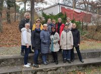 Eine Gruppe von Menschen steht draußen auf mehreren Steinstufen zusammen. Sie tragen überwiegend herbstliche oder winterliche Kleidung wie Jacken und Schals. Im Hintergrund sind ein Gebäude, Bäume mit herabgefallenen Blättern und Sträucher zu sehen. Die Szene wirkt wie ein Gruppenfoto während einer Veranstaltung oder eines Kurses in herbstlicher Umgebung.