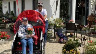 Ältere Frau mit Blumenstrauß sitzt vorne in einer roten Fahrradrikscha, diese wird von einer Frau mit Fahrradhelm gesteuert. 