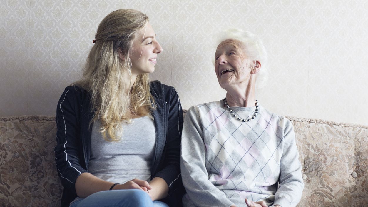 Junge Frau und Seniorin lächeln sich sitzend auf einem Sofa an.