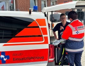 Zwei Personen in Malteser Einsatzkleidung stehen unter der geöffneten Heckklappe eines Einsatzfahrzeuges und sind im Gespräch.