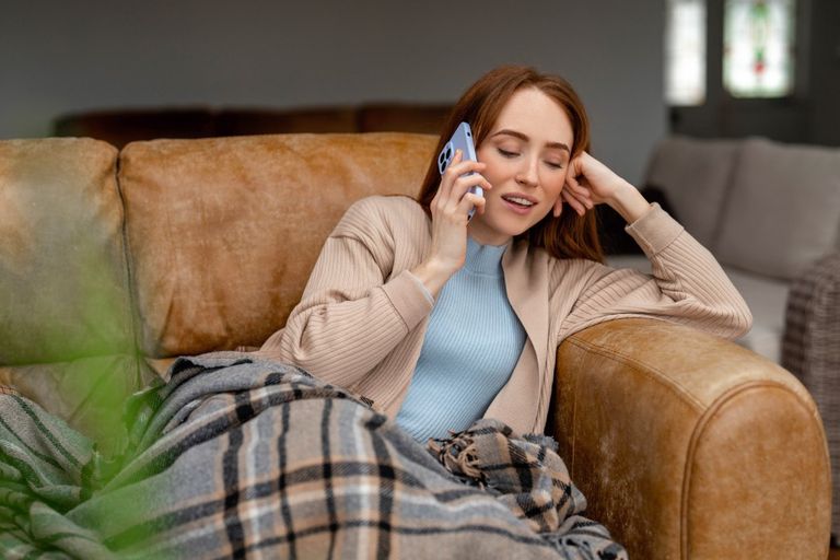Eine junge Frau sitzt mit einer Decke gemütlich auf einem Sofa und telefoniert mit einem Smartphone