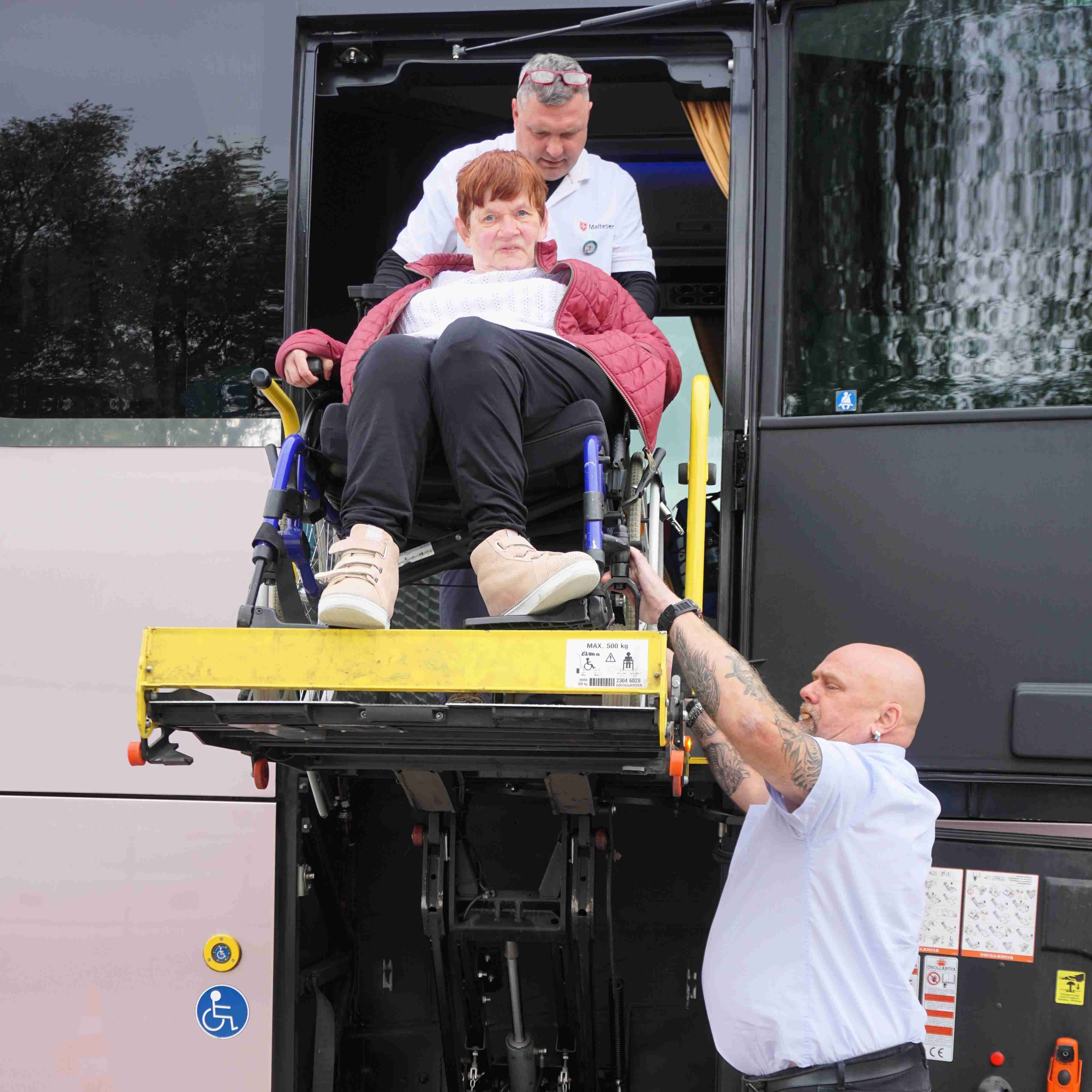 Person in Rollstuhl auf einer Hebebühne an einem Bus, begleitet von zwei Personen, eine mit weißem Hemd.