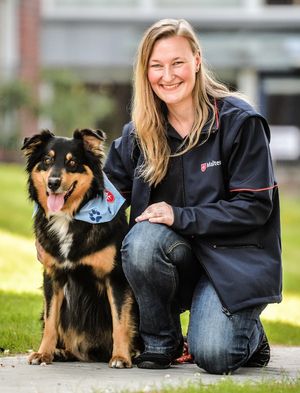 Lächelnde Frau kniet neben einem Hund, der ein Halstuch trägt.