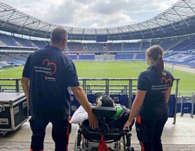 Torsten Richteweg im Rollstuhl mit den beiden Maltesern an der Seite auf den Rängen der Heinz von Heiden Arena