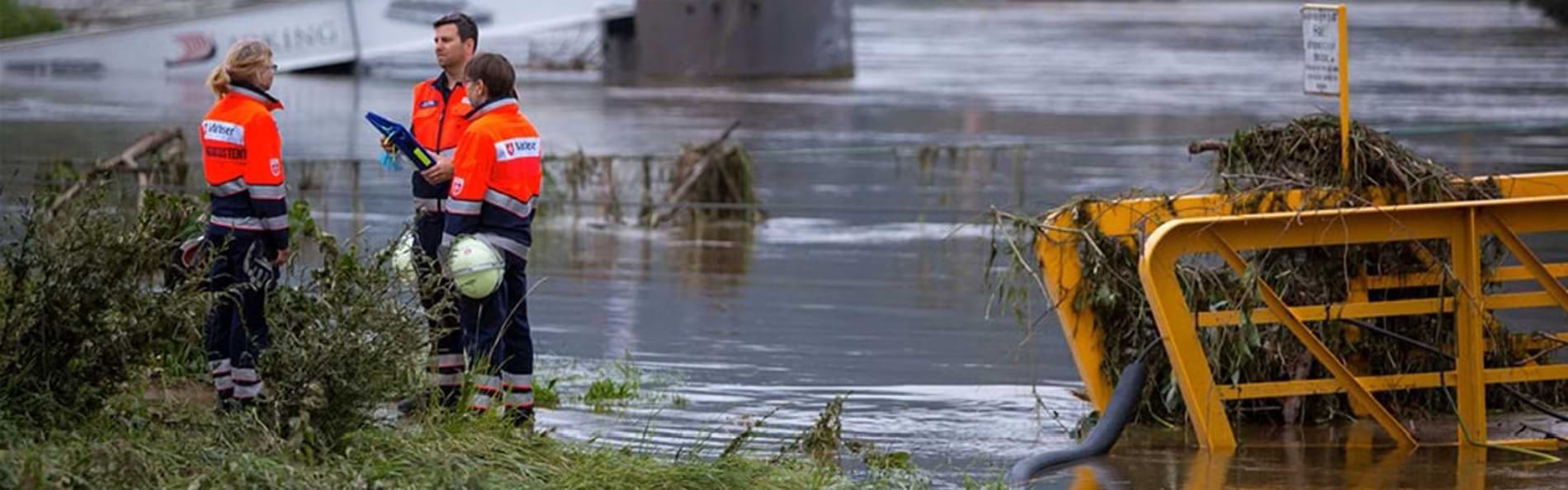 Drei Einsatzkräfte des Katastrophenschutzes stehen an einem überschwemmten Flussbett.
