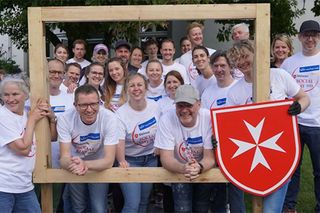 Menschen mit Firmentrikots blicken durch einen großen Bilderrahmen mit Malteserwappen