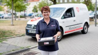 Mitarbeiterin des Malteser Menueservice trägt eine Warmhaltebox und lächelt in die Kamera.