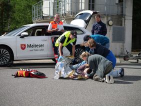 Ein Malteser PKW steht im Hintergrund mit geöffneter Heckklappe. Im Vordergrund üben 4 Jugendliche die Versorgung eines Verletzten. 2 Prüfer beobachten die Szene.