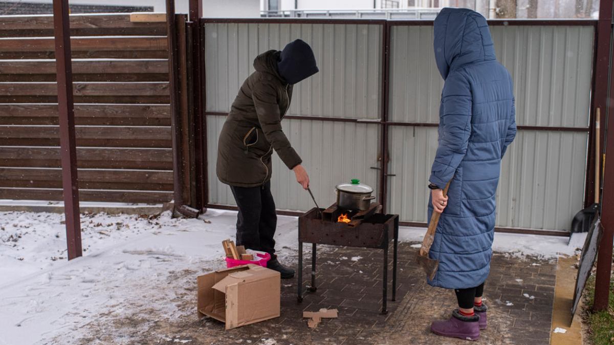 Zwei Menschen in Winterjacken kochen draußen etwas in einem Topf, der auf einem Grill steht.