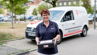 Mitarbeiterin des Malteser Menueservice trägt eine Warmhaltebox und lächelt in die Kamera.