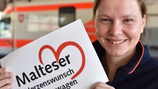 Malteser Mitarbeiterin zeigt das Logo des Herzenswunschkrankenwagens in die Kamera.