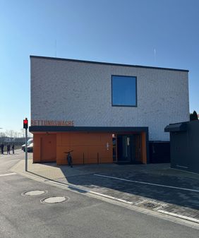 Ein modernes Gebäude mit einer hellen Klinkerfassade und einem großen rechteckigen Fenster im oberen Bereich. Im Erdgeschoss befindet sich ein orangefarbener Eingangsbereich mit der gut sichtbaren Beschriftung „Rettungswache“. Vor dem Gebäude steht ein Fahrrad. Links am Straßenrand ist eine rote Fußgängerampel zu sehen und im Hintergrund stehen einige Personen. Der Himmel ist klar und blau.