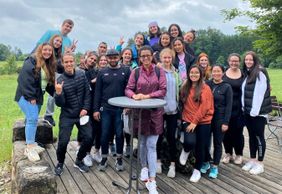 Gruppe junger Menschen posiert fröhlich im Grünen auf Holzterrasse vor bewölktem Himmel – gemeinsamer Ausflug oder Team-Event.