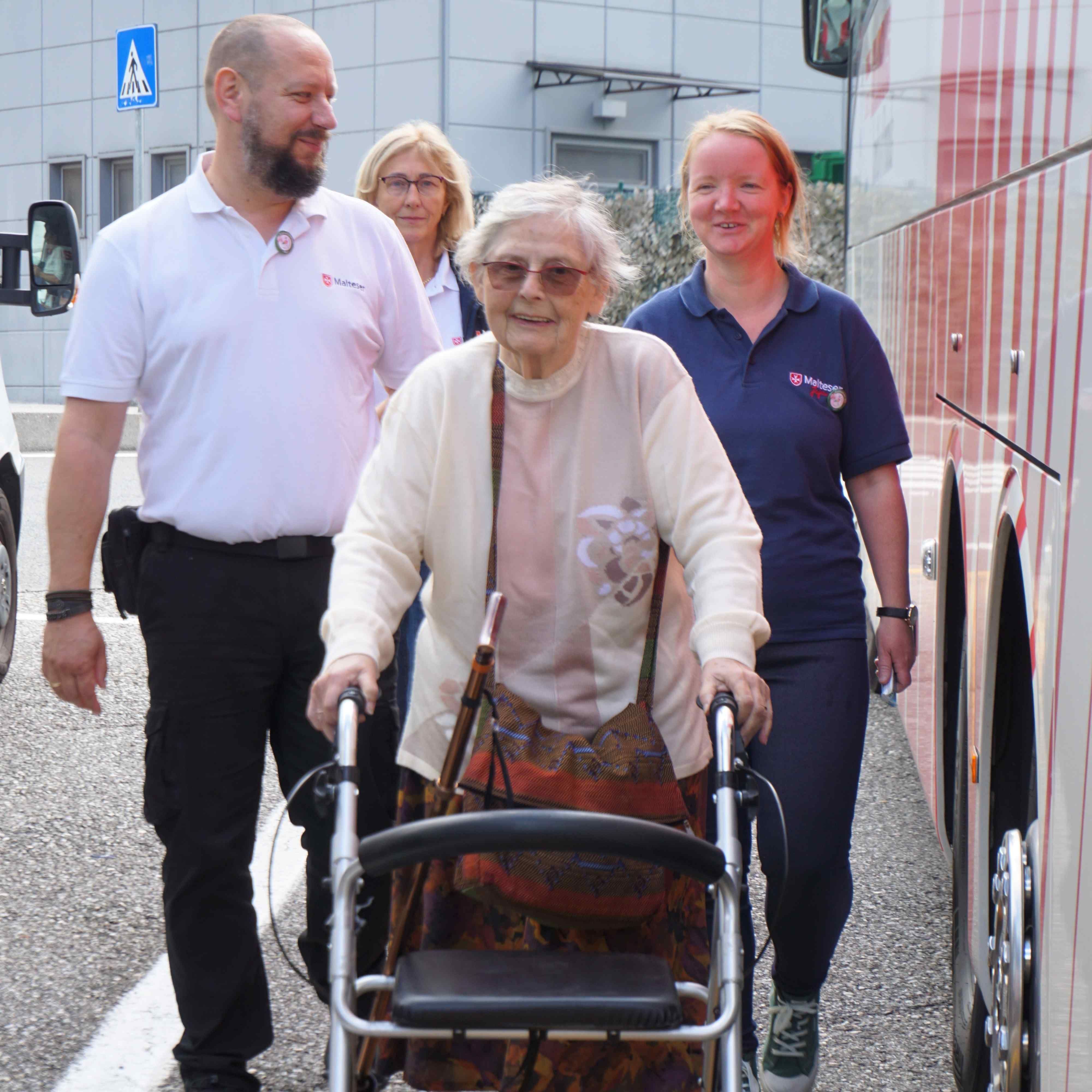 Ältere Frau mit Rollator wird von zwei Personen in Polohemden mit Logo begleitet, sie gehen neben einem Bus auf einem Gehweg.