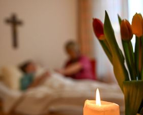 Eine Kerze im Vordergrund. Im Hintergrund eine Ehrenamtliche am Bett im Gespräch mit einer Frau.am Bett einer 