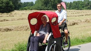 Ein Mann in Lederhose steuert eine Malteser Rikscha mit rotem Dach, vorne sitzt eine ältere Dame, im Hintergrund ein Getreidefeld