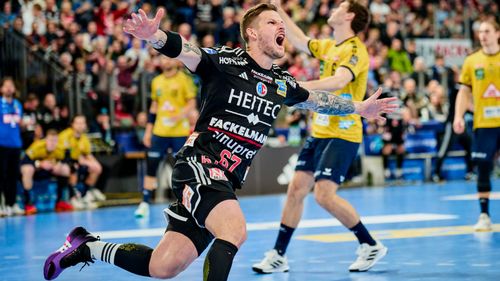 Handballspieler in Spielszene mit ausgestreckten Armen, weitere Spieler im Hintergrund sichtbar.