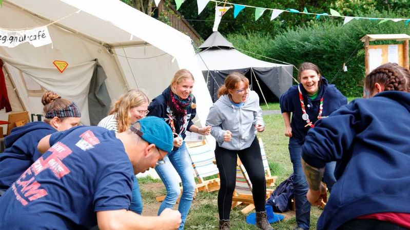 Eine Gruppe junger Menschen tanzt in einem Kreis, dahinter Zelte auf einer Wiese