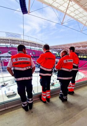 Vier Personen in orange-schwarzen Malteser-Sanitätsdienst-Westen blicken auf ein Stadion mit rotem Sitzbereich und Laufbahn.