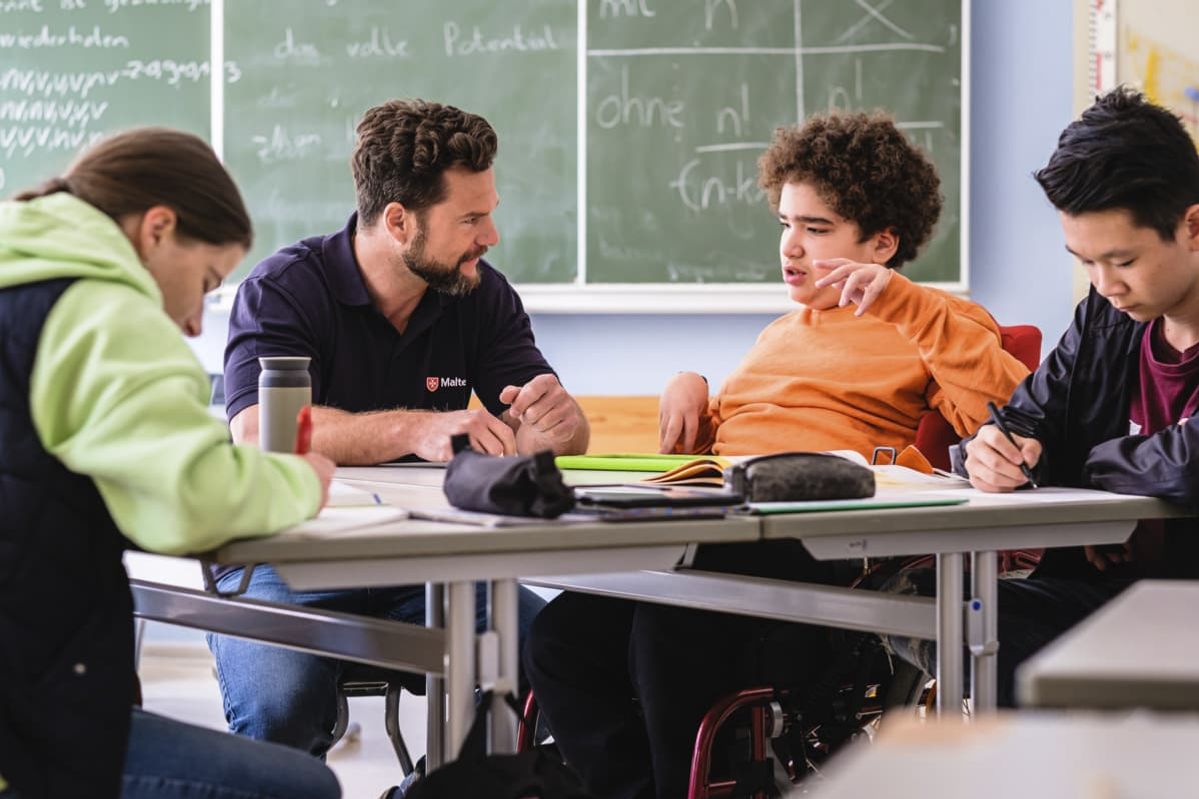 Malteser Schulbegleiter sitzt mit Schülern im Klassenzimmer.