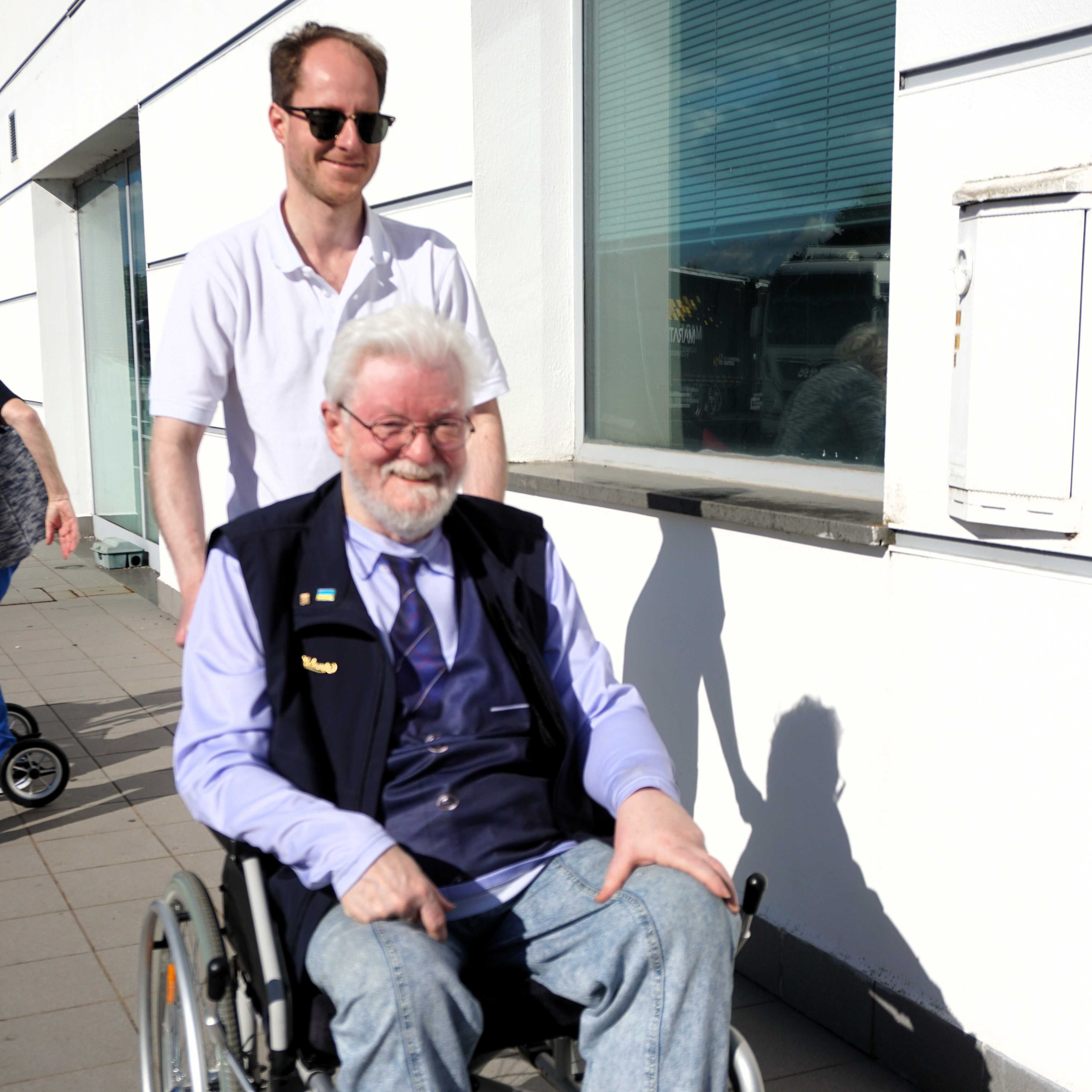 Älterer Mann mit weißem Haar sitzt im Rollstuhl, trägt blaue Jeans, hellblaues Hemd und dunkle Weste. Junger Mann in weißem Poloshirt schiebt den Rollstuhl auf einem Gehweg neben einem Gebäude mit großem Fenster.