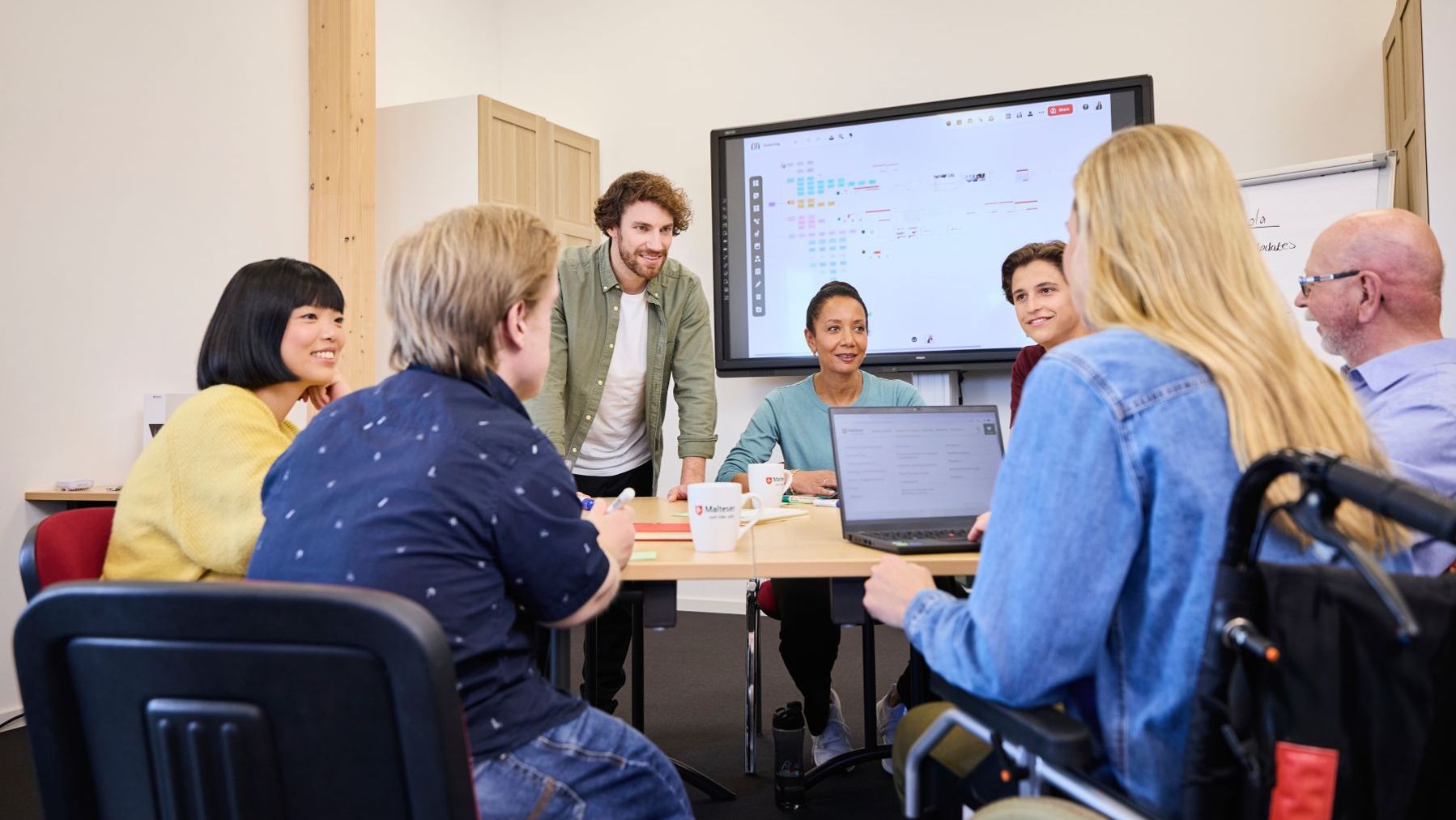 Gruppe von Maltesern bespricht sich in einem Büro, im Hintergrund ein Bildschirm