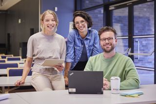 Drei junge Menschen – zwei Frauen und ein Mann – befinden sich in einem Schulungsraum der Malteser. Der Mann sitzt vor einem geöffneten Laptop am Tisch und die beiden Frauen sitzen links hinter ihm auf der Tischplatte. Alle drei lächeln in die Kamer