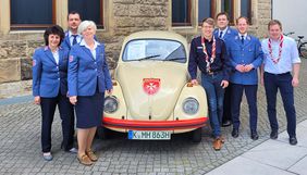 Mehrere Frauen und Männer stehen links und rechts von einem alten VW Käfer.