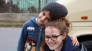 Junge Frau in Malteser Jacke mit einem Glücksbringer Paket wird von einem Kleinkind mit Mütze und bunter Strickjacke umarmt, im Hintergrund steht ein Malteser Fahrzeug